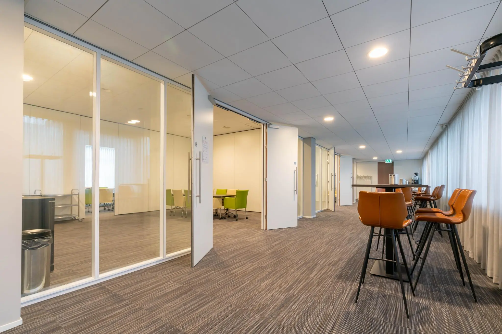 Moderne vergaderruimte aan de De Bruyn Kopsstraat met glazen wanden, open deuren en een zitgedeelte met hoge stoelen.